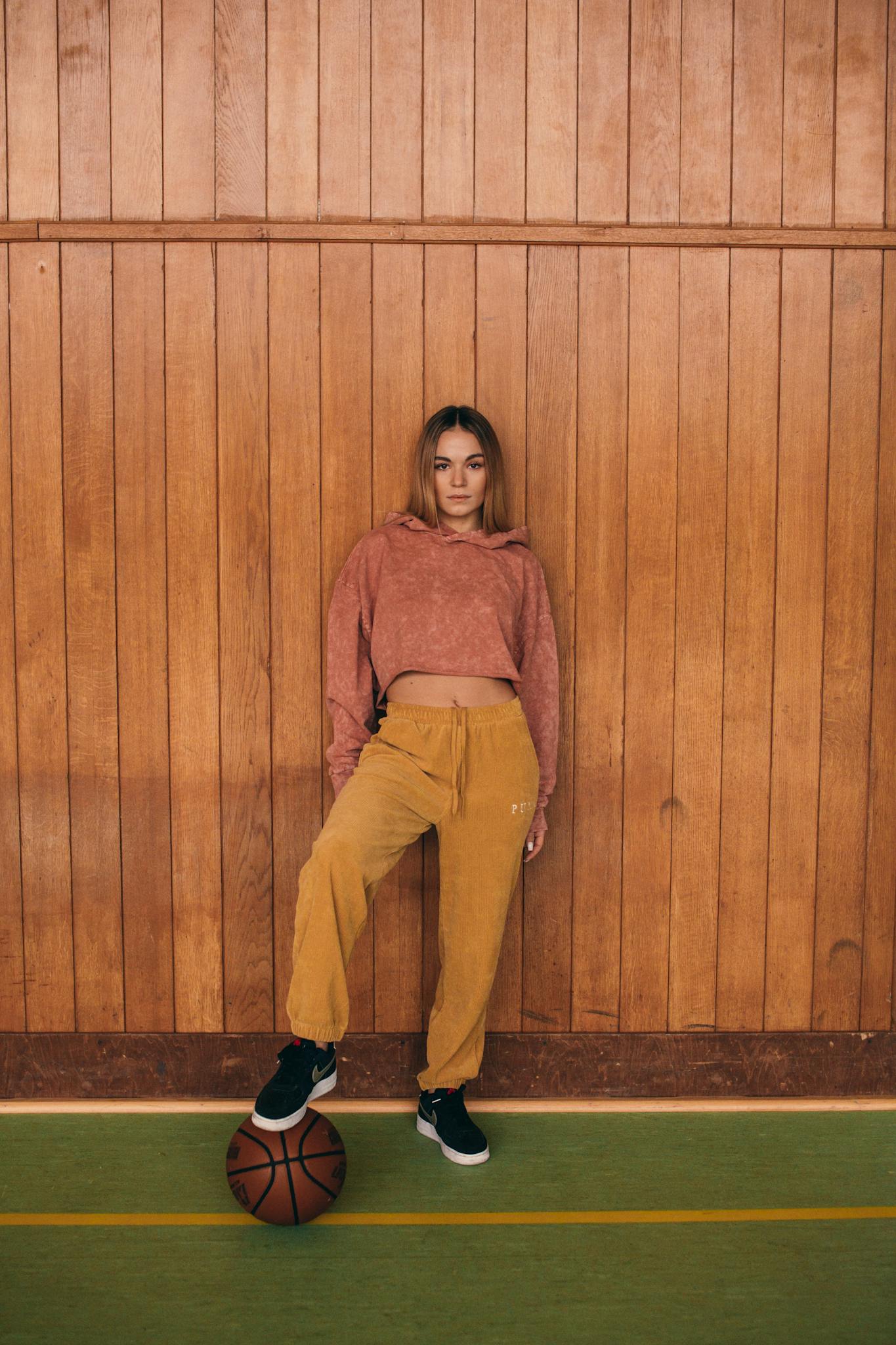 A young woman leans against a wooden wall with a basketball at her feet, wearing casual sportswear.